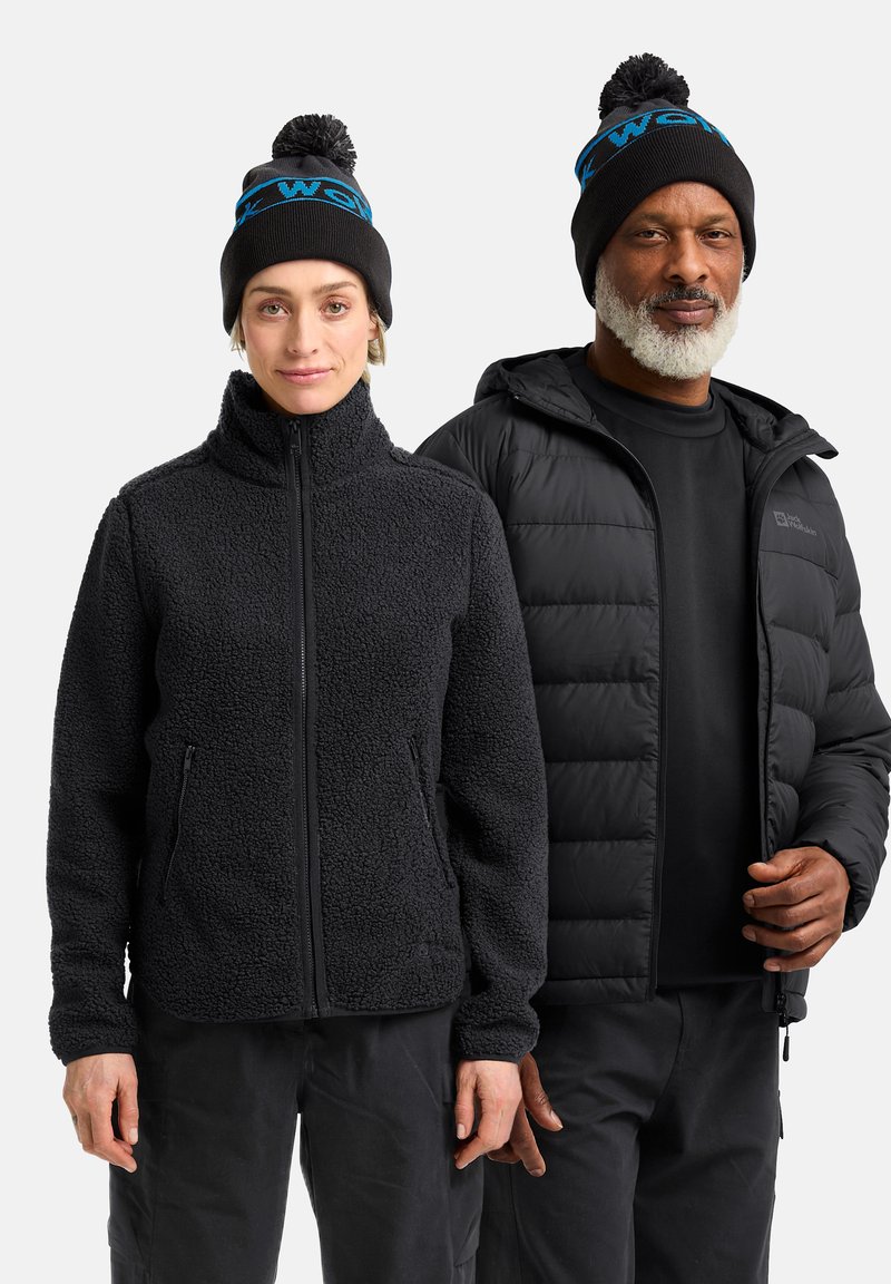 Giacca di pile nera con colletto alto e tasche con zip su una donna; uomo in una giacca imbottita nera, entrambi indossano berretti di lana neri e blu.
