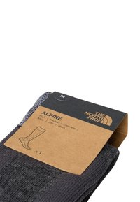 Sive pletene nogavice z rebrasto teksturo, opremljene s kartonsko oznako z napisom "ALPINE" in velikostjo "M". Namenjene za intenzivno nošenje.