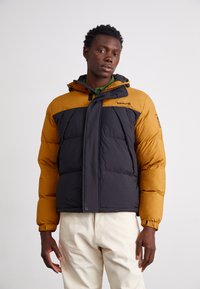 Pufferjakke med en guldbrun overdel og sort krop, med elastiske ærmer, hætte og en frontknaplukning. Materialet er glat og quilted.