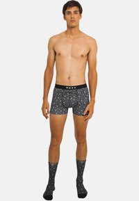 Boxer shorts noirs avec un motif blanc, design ajusté et une ceinture noire étiquetée "WAXX." Associés à des chaussettes noires à motif assorti.