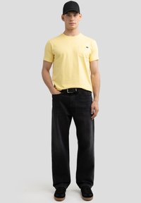 Geltona medvilninė marškinėliai su krūtinės kišeniu, juodi džinsai ir juodi sportbačiai su rudais padu, dėvimi su juoda kepure.