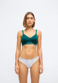 Tirkizni bralette s podesivim naramenicama, s suptilnim V-izrezom i elastičnom trakom s Calvin Klein logom. Uparen s bijelim gaćicama.