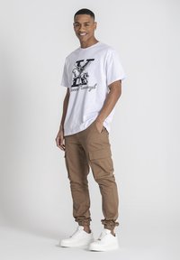 Camiseta blanca con estampado gráfico negro, combinada con pantalones cargo color marrón claro que tienen bolsillos con cremallera y puños elásticos, llevados con zapatillas blancas.