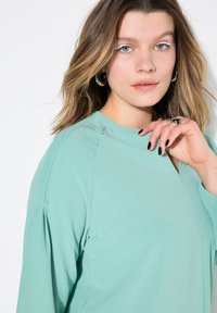 Top verde menta con maniche lunghe, texture morbida e scollo rotondo. Presenta leggeri plissettamenti sulle spalle e una vestibilità rilassata.