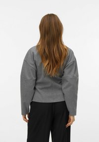 Šedá strukturovaná bunda s texturovaným povrchem, širokými rukávy a zkráceným střihem, nošená s volnými černými kalhotami. Zobrazený zadní pohled.