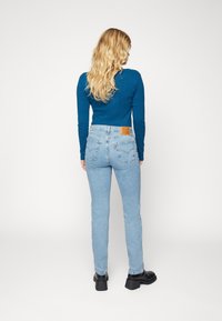 Jeans de mezclilla azul claro con un diseño ajustado, cintura alta y dos bolsillos traseros. Combinados con una blusa azul de manga larga y zapatos de tacón bloque negro.