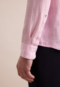 Camicia di lino rosa chiaro con polsini a bottoni, caratterizzata da un piccolo dettaglio ricamato sulla manica e una finitura in tessuto testurizzato.
