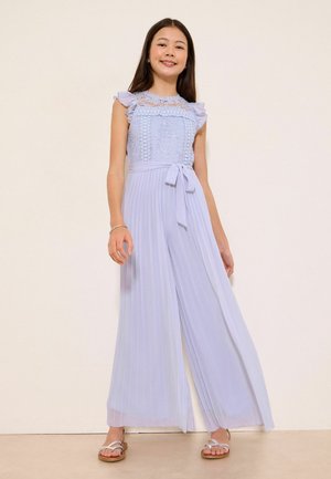 Młoda dziewczyna uśmiecha się, nosząc jasnoniebieski bezrękawnik z koronką z plisowanymi szerokimi spodniami i białymi sandałami.