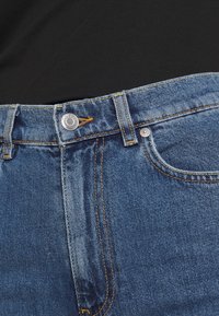 Gros plan de la ceinture d'un jean en denim bleu avec un bouton argenté et une boucle de ceinture sur un fond de tissu noir.