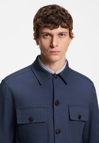 Tamsiai mėlyna striukė su struktūrizuotu apykakliu, dviem krūtinės kišenėmis ir juodais mygtukais. Lygi tekstūra su pritaikytu siluetu.