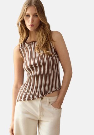 Vrouw met lang haar, die een bruin-wit gestreept mouwloos top draagt en lichtbeige broek met één hand in de zak.