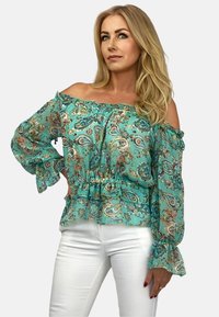 Turkusowa bluzka na jedno ramię z lekkiego materiału z kwiatowym wzorem paisley, falbaniastymi rękawami i elastycznym pasem. Połączona z białymi dżinsami.