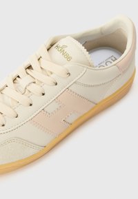 Bēša ādas sporta apavi ar gaiši rozā akcentiem, apaļu purngalu un plakanu gumijas zoli. Iekļautas teksturētas sānu paneļa un šņorēšanas detaļas. Zelta logo uz mēles.