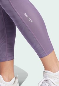 Fioletowe legginsy sportowe o dopasowanym kroju, z teksturowanymi wzorami, płaskimi szwami i białym logo na zewnętrznej stronie uda.