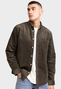 Camicia di velluto a coste color oliva scuro con colletto button-down, taschino sul petto, bottoni bianchi e maniche lunghe. Indossata sopra una maglietta bianca, abbinata a denim.