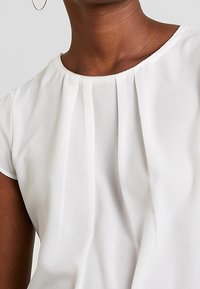 Vit blus med korta ärmar, rundad halsringning, subtila veck och lätt textur. Kanterna är snyggt avslutade, med en slät yta.