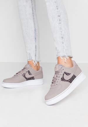 Persoon draagt lichtgrijze spijkerbroek met rafelige zomen en grijze sneakers met zwarte glitteraccenten en witte zolen, staand op een witte vloer.