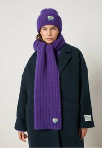 Vijolična kapa in šal iz mehkega, teksturiranega materiala. Šal ima navpične rebraste vzorce in logo blazinico. Pod njim je oblečen temen plašč.