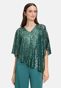 Top verde con paillettes, scollatura a V, maniche ampie e orlo asimmetrico, abbinato a pantaloni teal. Superficie lucida e con texture.