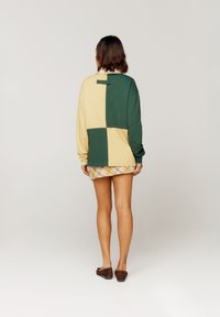 Sudadera color bloque verde y beige con mangas largas y cuello redondo, que presenta una pequeña etiqueta en la parte trasera y se lleva sobre una falda corta de cuadros.