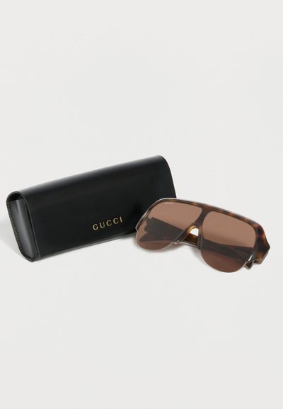 Lunettes de soleil écran oversize en écaille avec verres marron à côté d’un étui Gucci noir avec logo doré sur fond blanc.