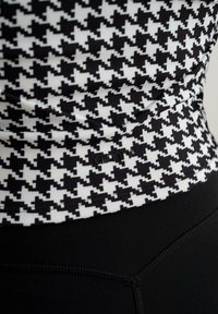 Juodas ir baltas houndstooth rašto viršus su juodu juosmeniu. Minkšta medžiaga su lygiu paviršiumi, šone turi diskretinį logotipą.