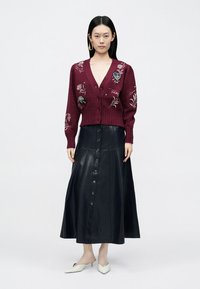 Bordowy dzianinowy kardigan z kwiatową haftowaną dekoracją, zestawiony z długą czarną skórzaną spódnicą z przodu z guzikami i rozszerzanym fasonem.