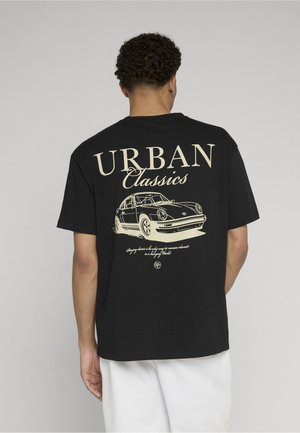 Mand iført sort t-shirt med vintage bilgrafik og teksten "Urban Classics" på ryggen, kombineret med hvide bukser, vendt væk.