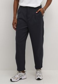 Pantalon bleu marine sur mesure avec une coupe décontractée, doté de deux plis avant et de poches latérales, associé à des baskets en maille argentée.