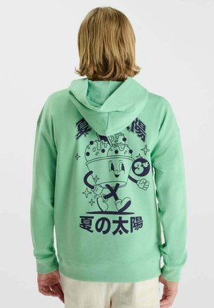 Persoon met halflang haar die een lichtgroene hoodie draagt met een cartoon ijsbeker met lopende benen en Japanse tekst op de achterkant.
