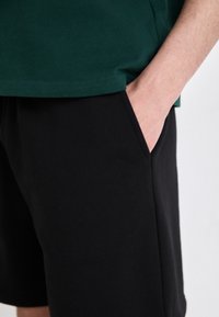 Pantalones cortos negros en una tela suave, con bolsillos laterales. Combinados con una camiseta verde, visible en la cintura con un dobladillo limpio y cosido.