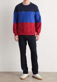 Sudadera con rayas horizontales en azul marino, azul real y rojo. Cuello redondo, mangas largas, mezcla de algodón. Combinada con pantalones negros y zapatillas blancas.