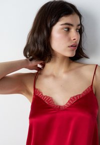 Camicetta in satin rosso con bordo in pizzo lungo il collo a V e spalline sottili e regolabili. Texture liscia e design aderente.