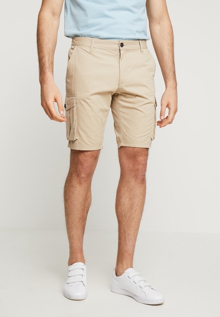 Beige cargo šortky vyrobené z lehkého materiálu, s bočními kapsami, předním zapínáním na knoflík a volným střihem. Nosené s bílými teniskami.