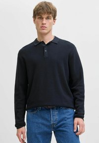 Joven con cabello corto castaño claro, vistiendo una camisa polo negra de manga larga y jeans azules, de pie contra un fondo liso claro.