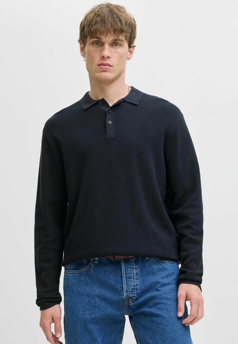 Joven con cabello corto castaño claro, vistiendo una camisa polo negra de manga larga y jeans azules, de pie contra un fondo liso claro.