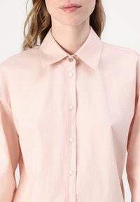 Light pink button-up shirt with a classic collar, featuring white buttons, a subtle texture, and a slightly relaxed fit.