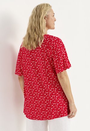 Frau mit langen blonden Haaren trägt ein rotes Kurzarmshirt mit kleinem weißem Blumenmuster und weiße Hosen, von hinten gezeigt.