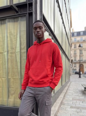 Jeune homme portant un sweat à capuche rouge et un pantalon gris, debout, une main dans la poche, à côté d’un bâtiment en verre et en métal dans une rue de la ville.
