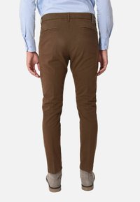 Pantaloni marroni slim-fit in cotone, con tasche posteriori e texture liscia, abbinati a stivali alla caviglia grigi. Polsino della camicia visibile.