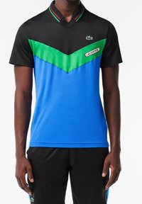 Man med kortärmad Lacoste sporttröja med svarta, gröna och blå färgblock, samt svarta shorts med blå sidostråk.