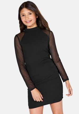 Noor tüdruk naeratab, kandes musta liibuvat kleiti läbipaistvate pikkade varrukate ja kõrge kaelusega, seistes tavalise heleda tausta ees.