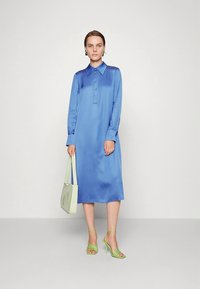 Vestido camisa de manga comprida em cetim azul saturado com decote de gola, fecho de botões e comprimento midi. Usado com acessórios verde claro.