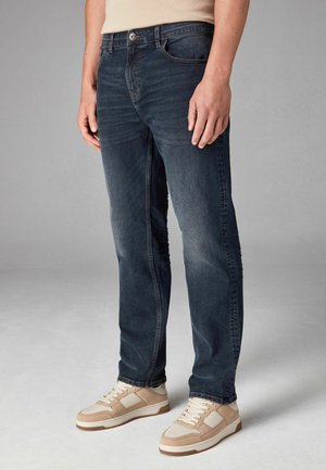 Vaqueros de mezclilla azul oscuro con corte recto, diseño de cinco bolsillos, detalles de costura sutiles y textura desgastada. Llevados con zapatillas beige.