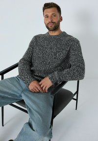 Pulover tricotat texturat, gri și negru, cu guler rotund, asociat cu blugi albaștri deschiși. Modelul stă pe un scaun negru, cu un fundal simplu.