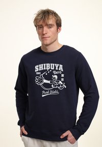 Sudadera azul marino con un estampado gráfico de un jugador de béisbol y el texto "SHIBUYA 1961 TOKYO Park Fields", con puños y dobladillo acanalados.