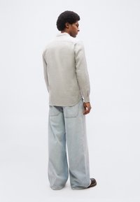 Άνδρας με αφροκτένισμα, φοράει ανοιχτό γκρι μπλούζα με μακριά μανίκια, φαρδιά ανοιχτό μπλε τζιν και σκούρα παπούτσια, στέκεται και κοιτάει πλάγια.