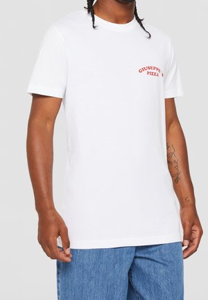 Mężczyzna w białej koszulce z krótkim rękawem z czerwonym napisem „Giuseppe's Pizza” na piersi oraz niebieskich dżinsach na jednolitym tle.