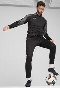 Svart treningsdress med grå detaljer, lange ermer, halvmaskin, og tettsittende bukser. Sorte fotballsko på en fargerik fotball.
