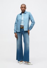 Light blue button-up shirt, light blue tank top, high-waisted flared denim jeans, and white heeled shoes, set against a plain background.